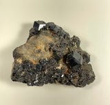 Dark gray to black garnet crystals with sharp, geometric faces clustered on a rough, earthy brown matrix against a neutral background.