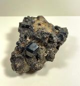 Dark metallic melanite garnet crystals with shiny cubic shapes clustered on a rough, gray-brown rocky matrix against a light neutral background.