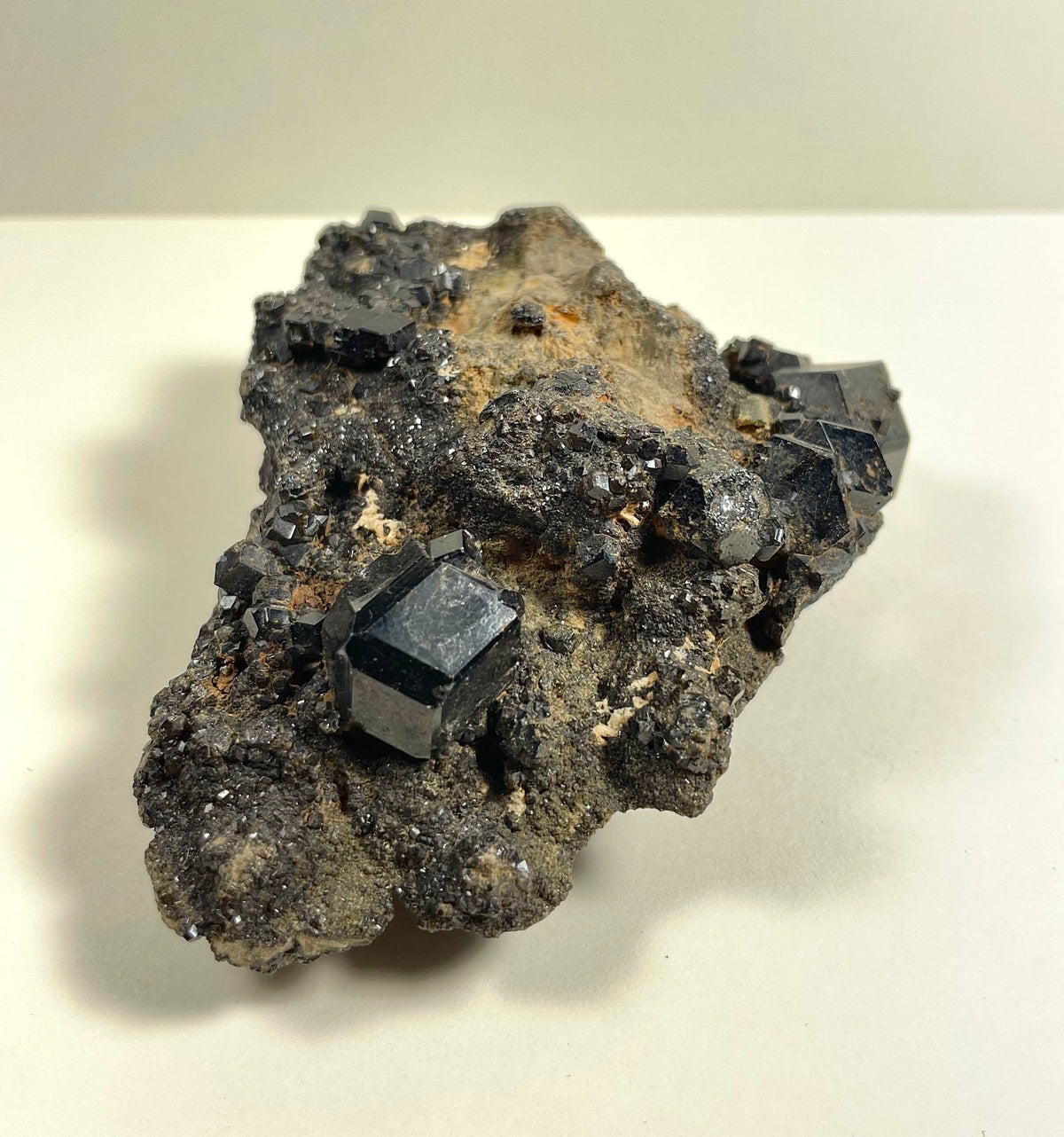 Dark metallic melanite garnet crystals with shiny cubic shapes clustered on a rough, gray-brown rocky matrix against a light neutral background.
