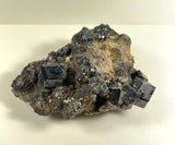 Dark metallic melanite garnet crystals with dodecahedral shapes clustered on rough brownish rock matrix with small glittering points, photographed on a light background.