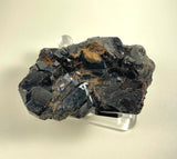 Dark black melanite garnet cluster with shiny, faceted crystal surfaces and patches of rusty brown matrix, displayed on a clear stand against a pale background.