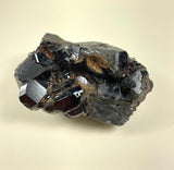 Dark metallic melanite garnet cluster with glossy, multifaceted crystal surfaces showing sharp edges and natural earthy inclusions on a neutral background.