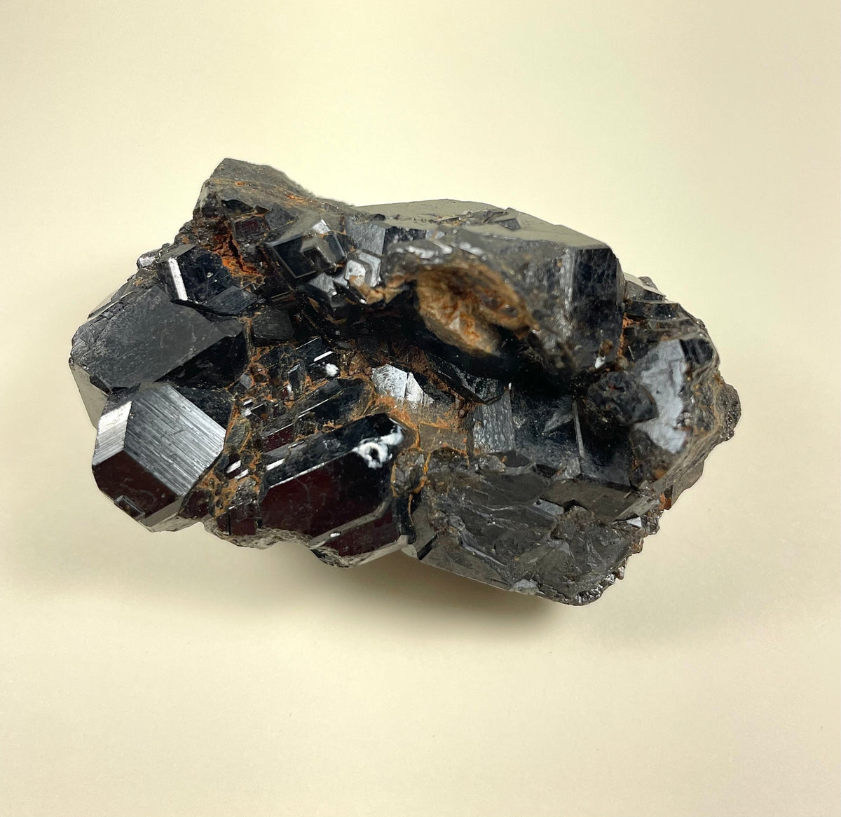 Dark metallic melanite garnet cluster with glossy, multifaceted crystal surfaces showing sharp edges and natural earthy inclusions on a neutral background.
