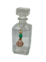 Clear rectangular glass decanter with textured surface, square stopper, and a jute string tied around its neck holding two wooden beads and a small scallop shell charm.