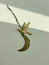 Brass ornament featuring a detailed bird in flight attached to a hammered crescent moon, hanging by a thin cord against a light background with diagonal shadow.