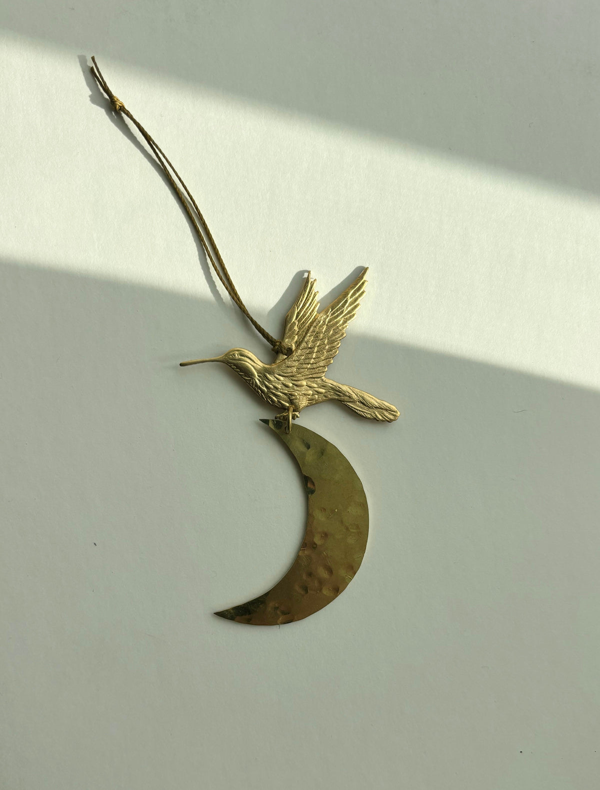 Brass ornament featuring a detailed bird in flight attached to a hammered crescent moon, hanging by a thin cord against a light background with diagonal shadow.