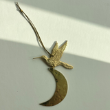 Over the Moon Ornament by Vida + Luz hanging against a white wall, showing slender crescent shape in hammered brass.