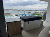 White rectangular RevX Acrylic X Tub Cold Plunge with black cover beside a matching beige slatted LUX chiller unit on a patio overlooking a marina under a cloudy sky.