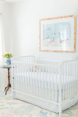 White baby crib with light blue bedding in a softly lit room, featuring a framed artwork of doves on a pale blue background above and a small wooden side table with a blue-and-white vase holding yellow tulips.