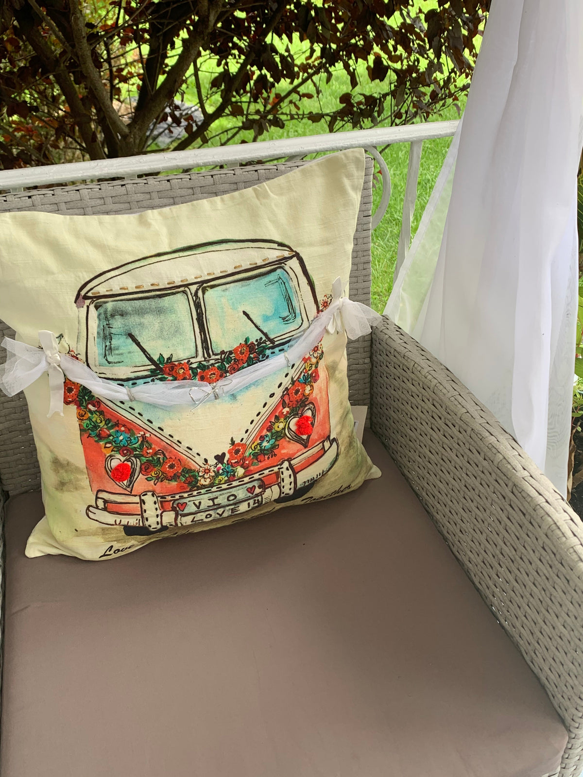 Square beige pillow with a colorful vintage van illustration adorned with red flowers and a banner reading 'Enjoy Love', placed on gray wicker outdoor chair with white sheer curtains.