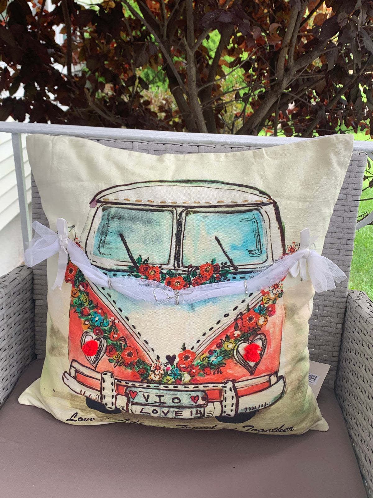 Cream throw pillow featuring a vintage van illustration decorated with colorful flowers and hearts, tied with a white ribbon at the front, set on a wicker chair.