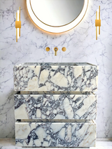 Freestanding vanity unit with a striking black and white marbled pattern, set against matching marbled wall tiles and flanked by two vertical gold sconces under a round mirror.