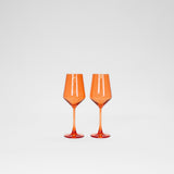 Two Aperol orange colored wine glasses with long stems and faceted bowls against a plain white background.