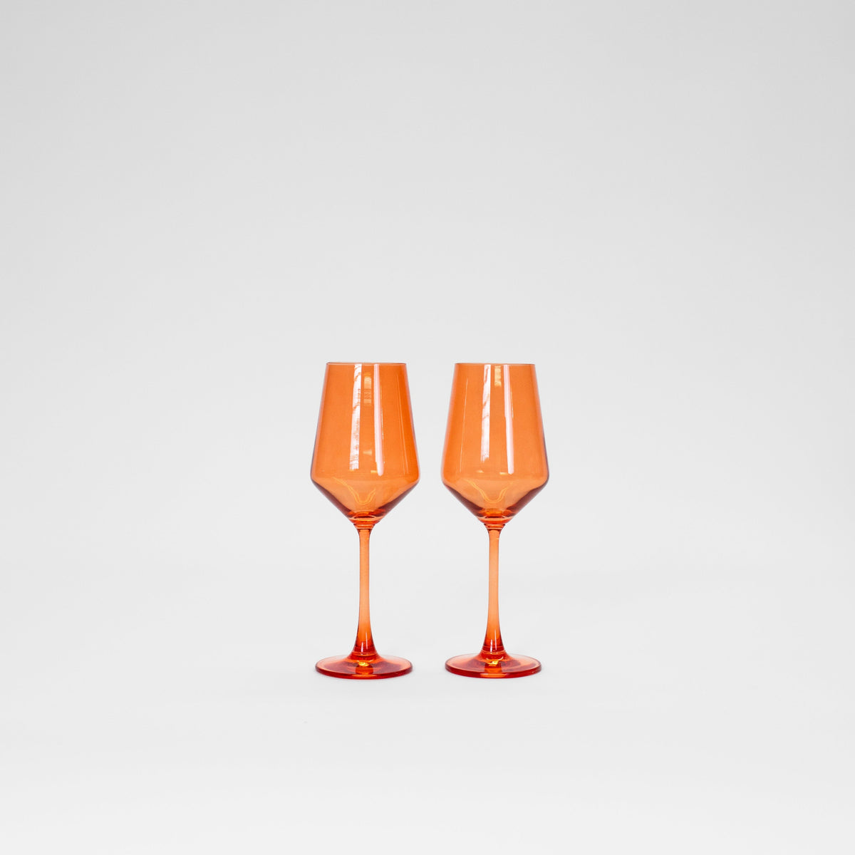Two Aperol orange colored wine glasses with long stems and faceted bowls against a plain white background.