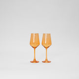 Two orange-tinted colored wine glasses with angular bowls and slender stems, set side by side against a plain white background.