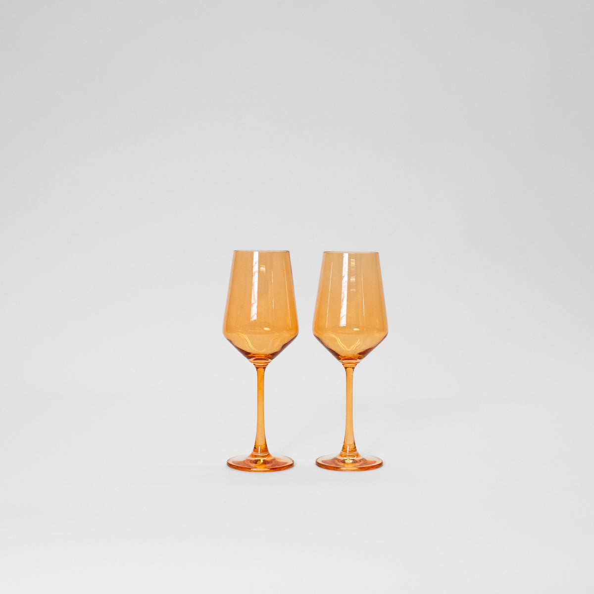 Two orange-tinted colored wine glasses with angular bowls and slender stems, set side by side against a plain white background.