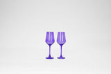 Two stemmed wine glasses in translucent veri peri purple, standing side by side against a white background with soft reflections on their surfaces.