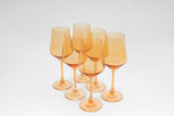 Six translucent orange stemmed wine glasses arranged in a loose cluster on a white background, showing their tall, slender stems and gently tapered bowls.