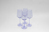 Four translucent pale lavender-colored wine glasses with long stems arranged in a diamond shape on a plain white background.