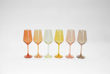 Six tall wine glasses in varying translucent colors—orange, peach, light green, yellow, pink, and smoky brown—arranged in a straight line on a white background.