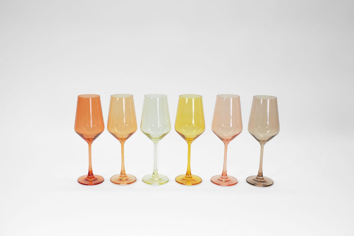 Six tall wine glasses in varying translucent colors—orange, peach, light green, yellow, pink, and smoky brown—arranged in a straight line on a white background.