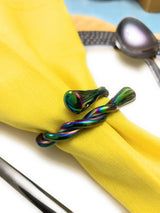 Two iridescent napkin rings with swirling tube and teardrop shapes, holding a bright yellow cloth napkin against a white background with silver utensils nearby.