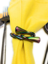 Iridescent rainbow-colored twisted metal napkin ring holding a folded yellow cloth napkin, with a stainless steel fork partially visible on the left.