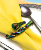 Iridescent rainbow-colored napkin ring with twisted and smooth shapes holding a yellow cloth napkin on a white plate with a spoon and fork nearby.