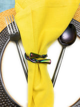 Iridescent rainbow-colored metal napkin ring twisted around a bright yellow napkin positioned on a white plate with a hammered dark gray rim, with matte black fork and spoon at sides.