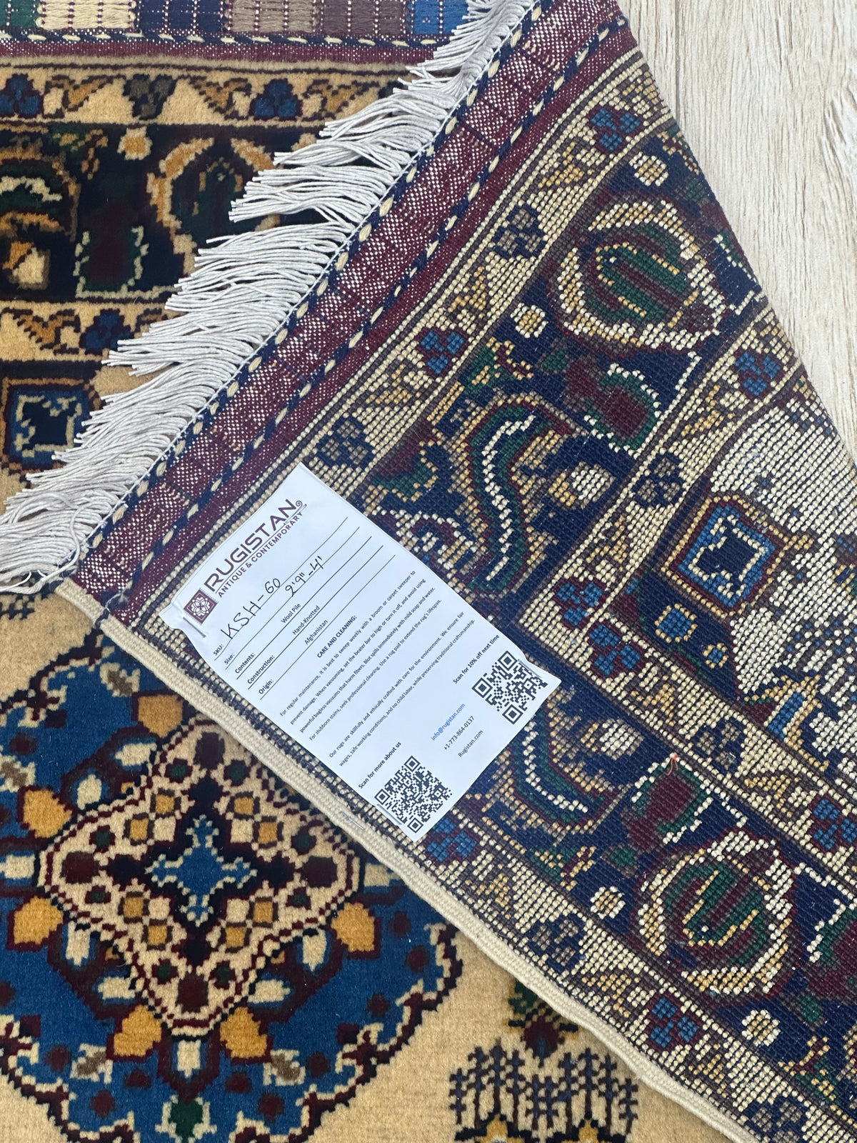 Corner of a hand-knotted Afghani Kashi wool rug with intricate geometric and floral patterns in blue, brown, red, and beige, showing fringe and a label from Rugistan.