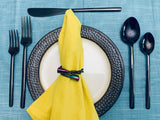 Matte black flatware set with two forks, a knife, a spoon, and a teaspoon arranged on blue fabric around a white plate with a textured dark metallic charger and a yellow napkin with a twisted metallic napkin ring.
