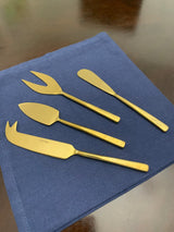 Set of four matte gold-toned cheese knives and spreaders with varied blade shapes, arranged on a folded dark blue cloth on a wooden surface.