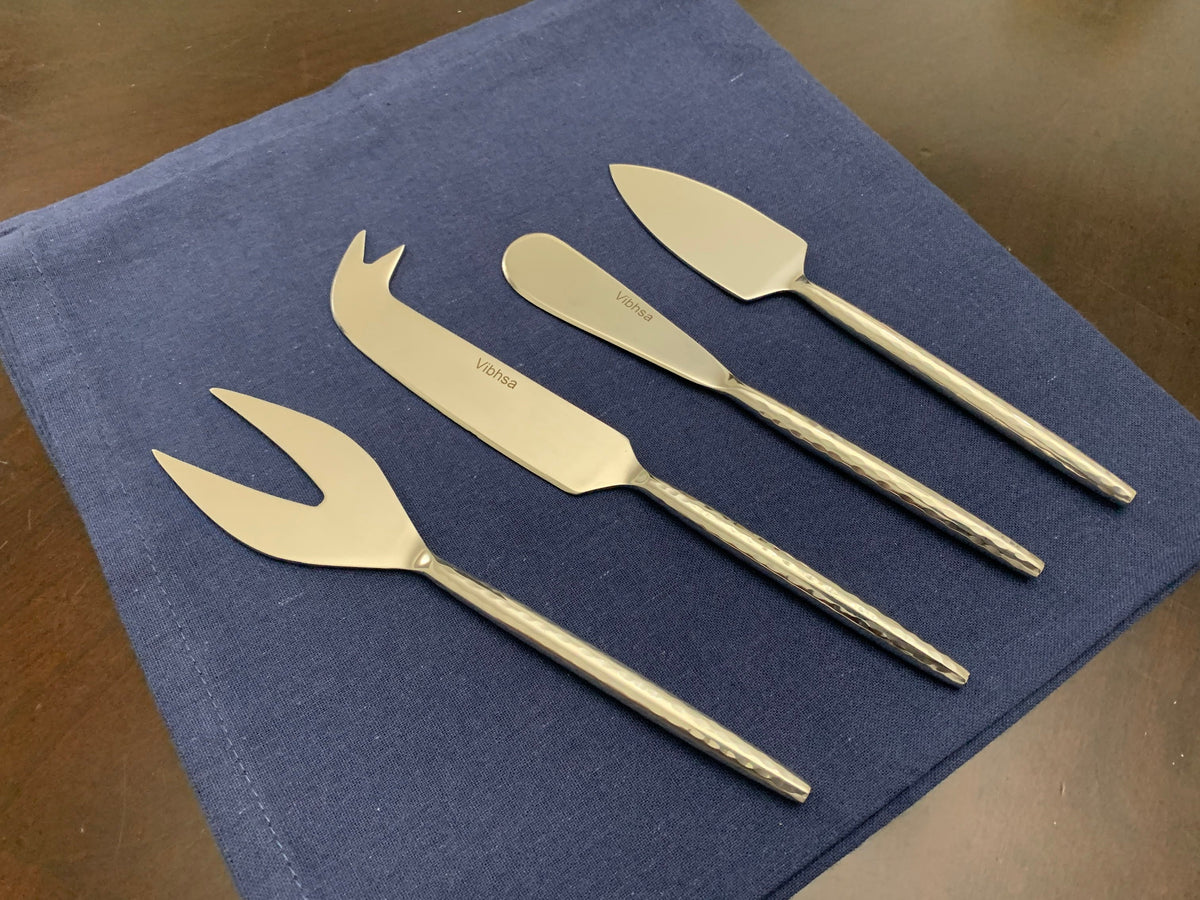 Set of four silver stainless steel cheese knives with textured handles displayed on a dark blue cloth, each knife head featuring a distinct shape for different cheese types.