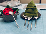 Hammered stainless steel flatware spoon resting on a pot with a red cloth rose, and four pieces leaning against a plate with a folded green napkin shaped like a Christmas tree on a blue tablecloth.