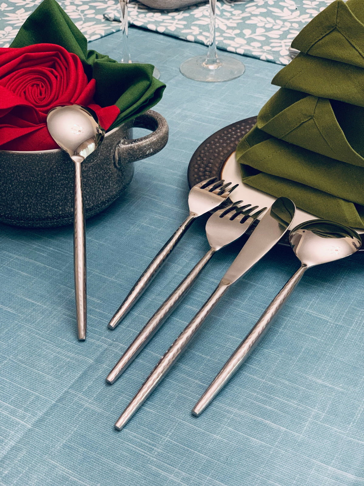 Hammered stainless steel flatware set including forks, spoons, and a ladle, arranged on a light blue tablecloth with folded green napkins and decorative red rose.
