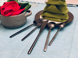 Matte black stainless steel fork, knife, teaspoon, tablespoon, and butter knife arranged beside a green fabric napkin folded into a triangular layered shape on a dark plate.