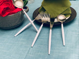 Set of white-handled stainless steel flatware including a spoon, two forks, a knife, and a ladle arranged on a blue tablecloth beside a folded green napkin on a dark plate and a red rose in a gray bowl.