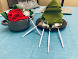 White-handled stainless steel flatware set arranged with green folded napkins on a light blue tablecloth, alongside a round gray pot with a red and green napkin inside.