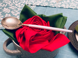White and silver stainless steel soup ladle resting diagonally on a red and green folded napkin inside a textured gray cup on a blue and white floral tablecloth.