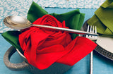 Silver spoon and fork placed over green and red cloth napkins folded into rose shapes, set inside a rustic gray ceramic bowl on a blue tablecloth.