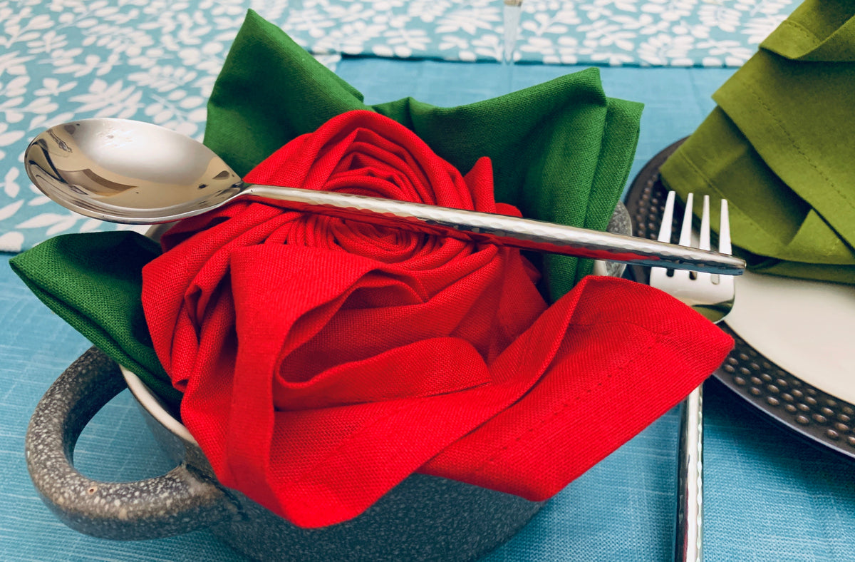 Silver spoon and fork placed over green and red cloth napkins folded into rose shapes, set inside a rustic gray ceramic bowl on a blue tablecloth.