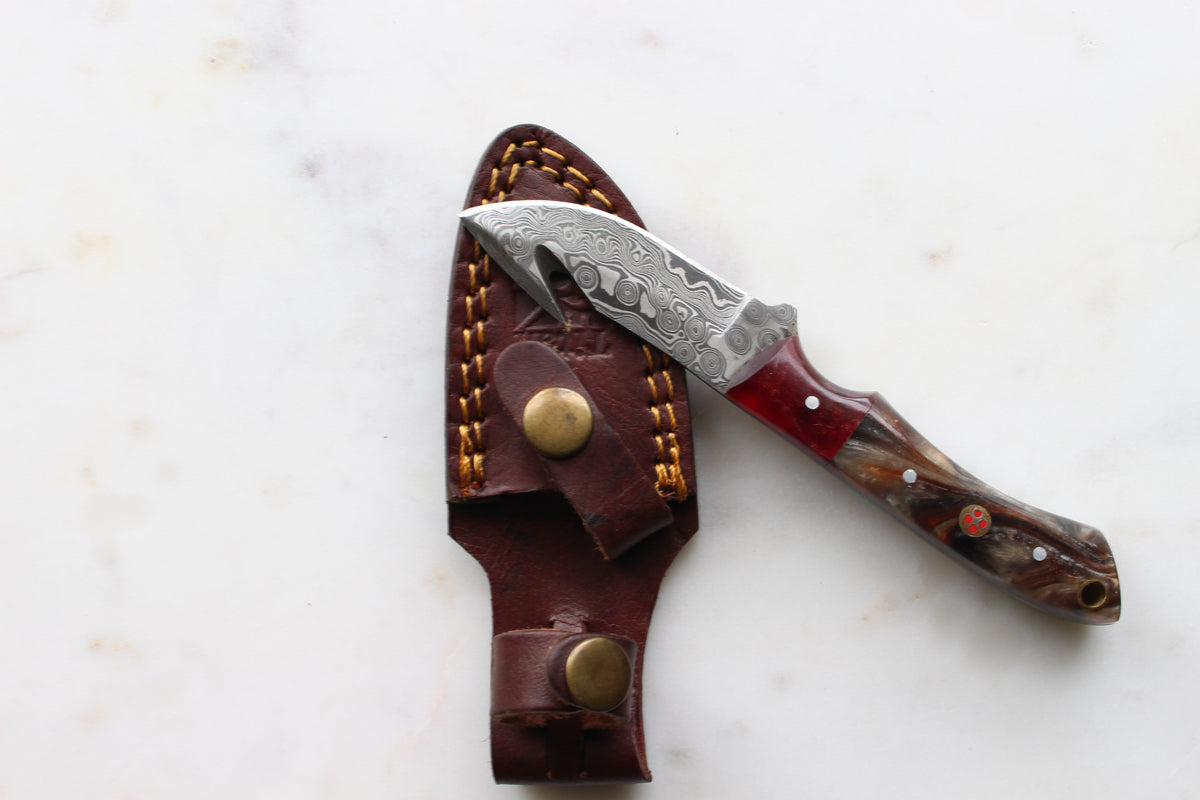 The Ravager Damascus gut hook hunting knife with red and brown resin handle resting on rough wood