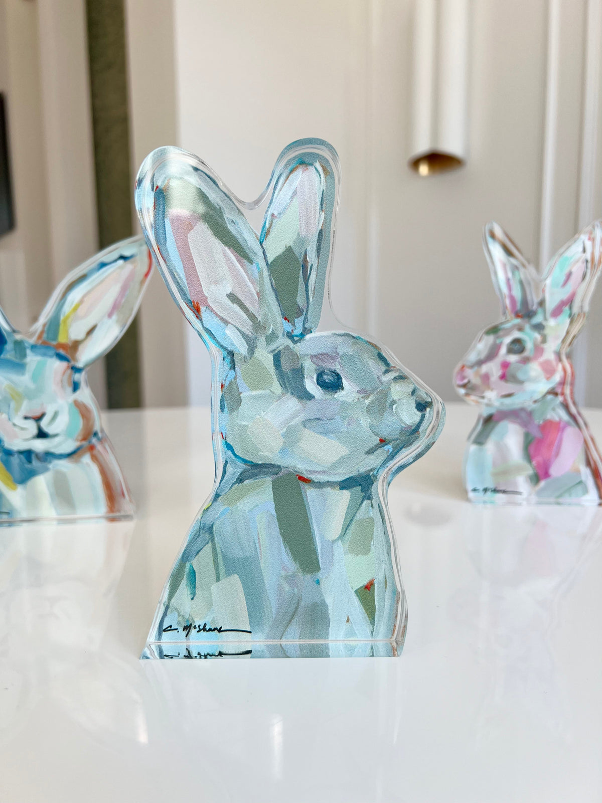Three translucent acrylic rabbit sculptures with pastel brushstroke patterns in blues, pinks, and greens, displayed on a glossy white surface.