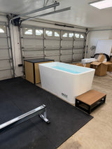 Rectangular white acrylic cold plunge tub filled with water, placed in a garage with black rubber floor mats and tan wooden step stool beside it.