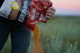 Person pouring orange-colored bait or feed from a red and white bag labeled 'Big Tine Triggered' into a green outdoor field during sunset.
