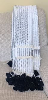 Off-white woven throw with black vertical stripes and large black tassels, draped over a light beige fabric couch arm.