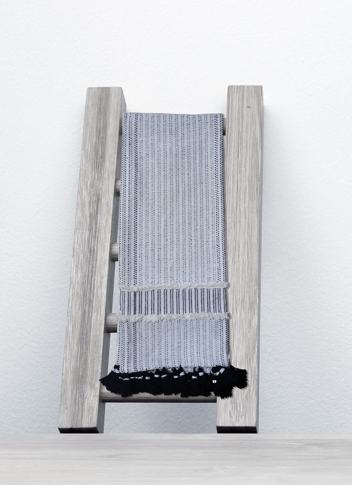 Light gray wooden ladder-style rack holding a folded light gray woven throw with black fringe against a white wall and light wood surface.