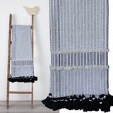 Hand loom woven textured throw in black and off-white with horizontal stripes and fringed edges, draped over a wooden ladder with a bird decoration on top.
