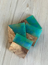 Four square coasters featuring a combination of natural brown wood grain and a blue-green epoxy resin with a swirling pattern, arranged overlapping on a light wood surface.