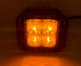 Square LED pod light with four amber-colored lenses illuminated inside a black heat sink casing, casting a warm yellow-orange glow on the surface below.